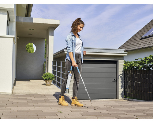 Kvinna tar bort ogräs med en gasbrännare på en stenlagd yta framför ett hus.