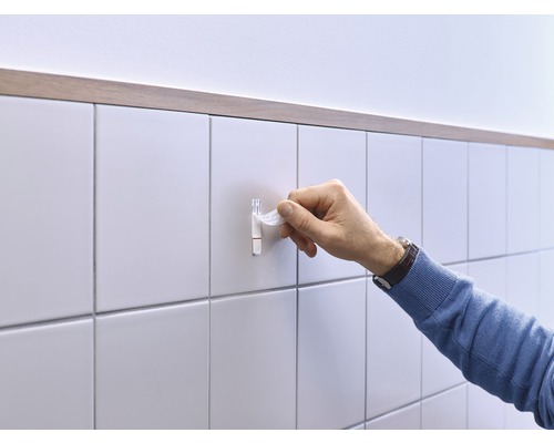 En hand fäster en självhäftande krok på en kakelvägg.