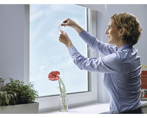 En kvinna fäster en fönsterkrok på ett fönster, krukväxter och dekorationer står på fönsterbrädan.