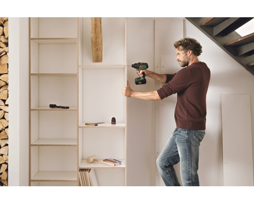 En man monterar en vit bokhylla med en sladdlös skruvdragare i ett rum med en stapel trä och en trappa.