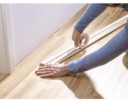En person fäster tejp på en golvlist på ett trägolv.