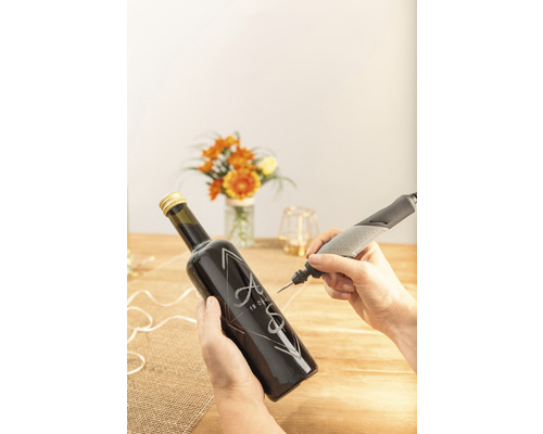 En person graverar en glasflaska med initialer och ett datum med ett Dremel-graveringsverktyg.