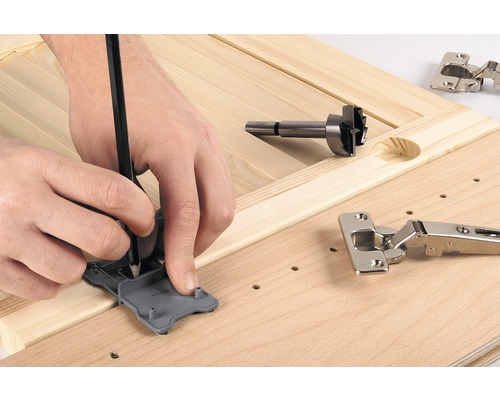 En person markerar positionen för ett gångjärn med en mall och en penna på en träskiva, bredvid en forstnerborr och ett gångjärn.