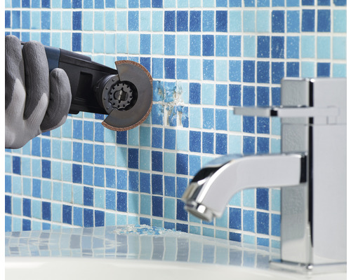 En arbetare med handskar tar bort kakel med ett multifunktionellt verktyg i ett badrum