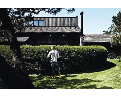 Man klipper häck med en häcksax i trädgården framför ett hus.