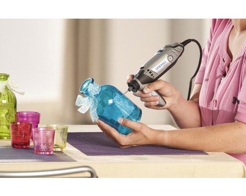 Kvinna graverar ett blått glas med ett Dremel multiverktyg. Dekorativa glasvaror i bakgrunden.