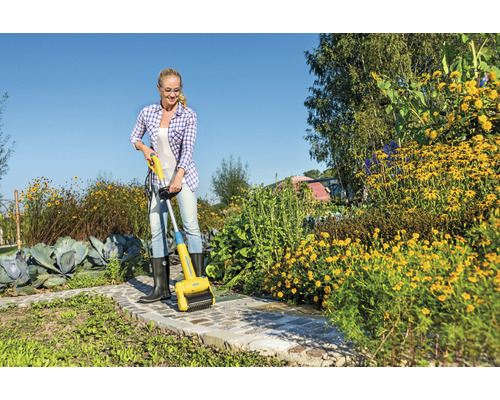 Kvinna tar bort ogräs med en elektrisk fogborste på en trädgårdsgång.
