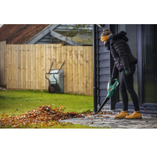 En kvinna använder en lövblås för att ta bort löv från en uteplats. I bakgrunden syns ett staket och en skottkärra.