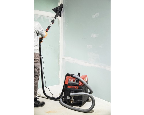 En person slipar en vägg med en gipsskivslipmaskin och en dammsugare.