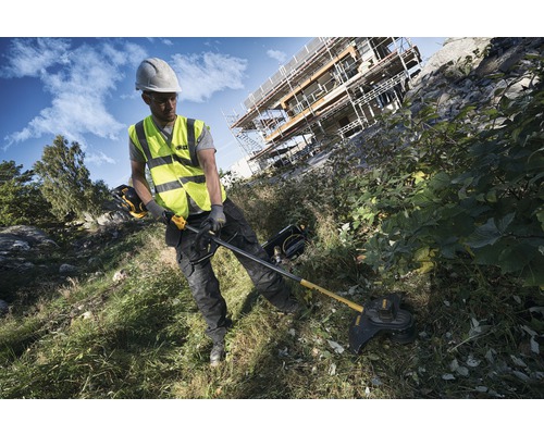 En byggnadsarbetare med skyddshjälm och säkerhetsväst använder en grästrimmer utomhus.