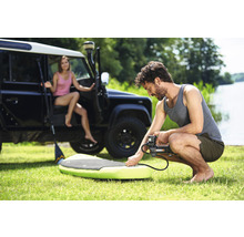 Man pumpar upp en stand-up paddleboard med en elektrisk pump, med en kvinna och ett terrängfordon i bakgrunden.
