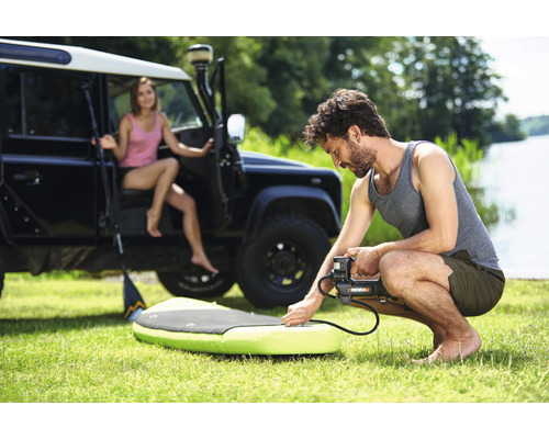 Man pumpar upp en stand-up paddleboard med en elektrisk pump, med en kvinna och ett terrängfordon i bakgrunden.