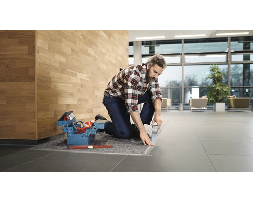 Hantverkare knäböjer på ett klinkergolv och arbetar på en övergångsprofil. Verktygslåda och vattenpass ligger redo.