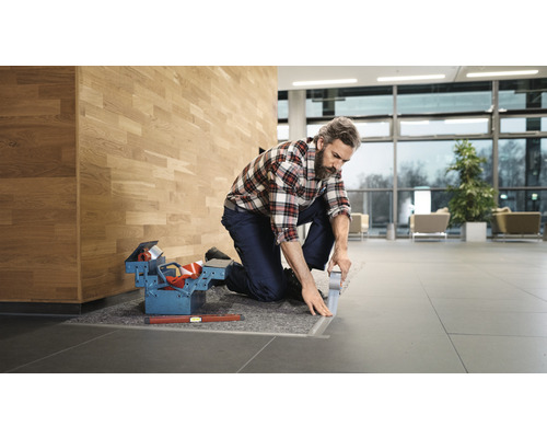 Hantverkare som knäböjer och limmar golvskenan med tejp, verktygslåda och vattenpass bredvid.