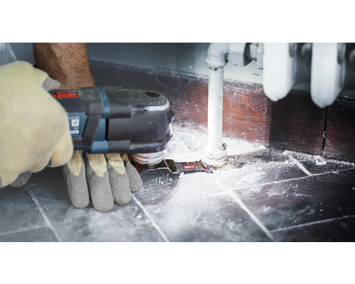 En person använder ett multifunktionellt verktyg på ett värmerör nära en vägg.