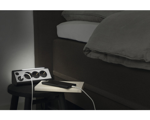 Grenuttag med USB-portar och en ljusramp på ett nattduksbord bredvid en säng.