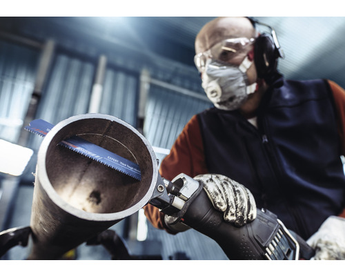 Man med skyddsglasögon, andningsskydd, hörselskydd och handskar sågar ett rör med en tigersåg.