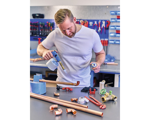 En man löder kopparrör med en gasbrännare och lödtenn på en arbetsbänk med verktyg och kopplingar.