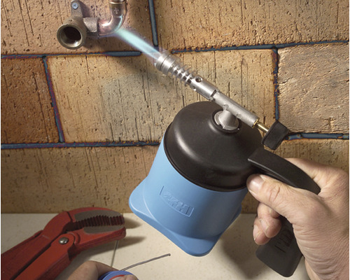 En hand använder en brännare för att löda rör på en tegelvägg.