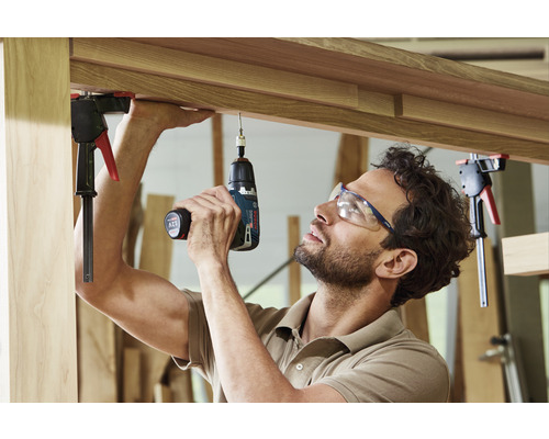 En man borrar med en sladdlös skruvdragare i ett arbetsstycke som är fastsatt med skruvtvingar.