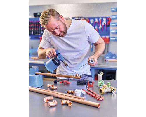 En man löder kopparrör med en brännare i en verkstad. På bordet ligger kopparrör, kopplingar och verktyg.