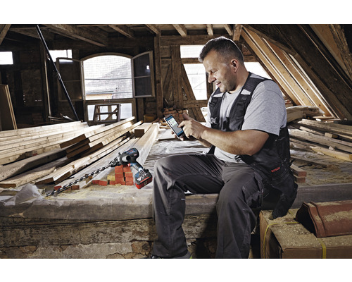 En man sitter på en vind och använder en mobiltelefon. Bredvid honom ligger en borrmaskin med en lång borr.