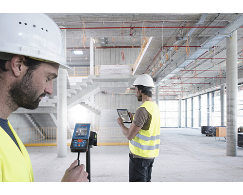 Två byggnadsarbetare använder en digital laseravståndsmätare och en surfplatta på en byggarbetsplats.