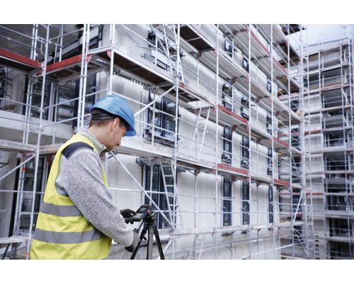 Byggnadsarbetare med hjälm och varselväst använder en laseravståndsmätare på ett stativ framför en byggnad med ställningar.