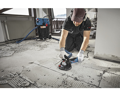 En man använder en betongslipmaskin på ett golv som är anslutet till en dammsugare. Bosch logotyp.