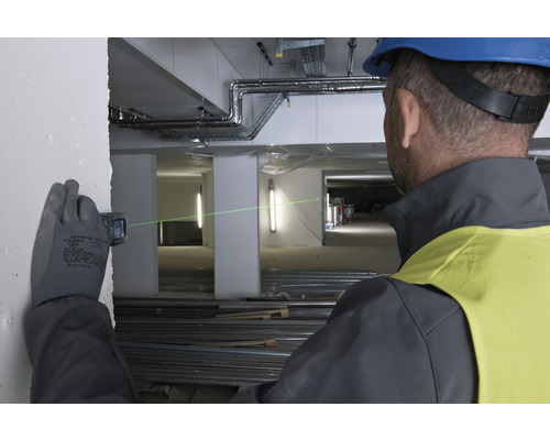 En man med skyddshjälm och handskar använder en laseravståndsmätare på en byggarbetsplats för att projicera en grön laserlinje på en vägg.