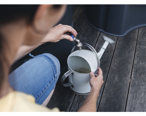 En person fyller en vit vattenkanna vid kranen i en trädgård.