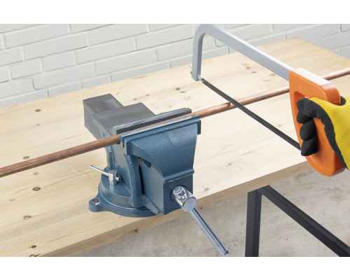 Ett skruvstäd håller ett kopparrör som skärs med en metallsåg.