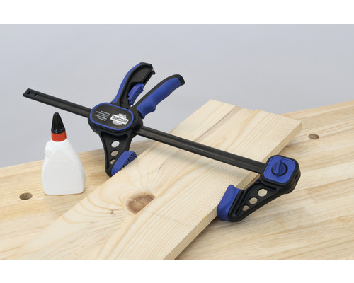 En enhandsspännare från INDUSTRIAL fixerar en träbräda på en arbetsbänk, bredvid en flaska trälim.