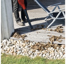 En person använder en lövblås för att ta bort löv från en träterrass. En solstol syns i bakgrunden.