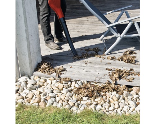 En person använder en lövblås för att ta bort löv från en träterrass. En solstol syns i bakgrunden.