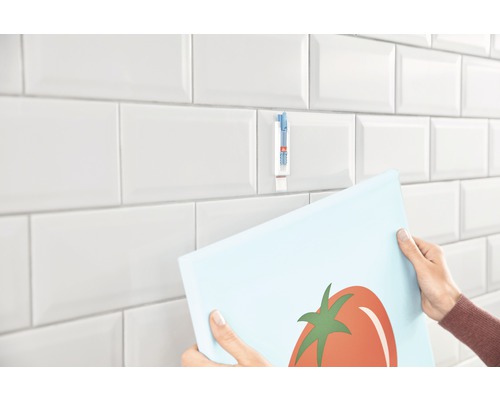 Person fäster en bild på en kakelvägg med en självhäftande remsa.