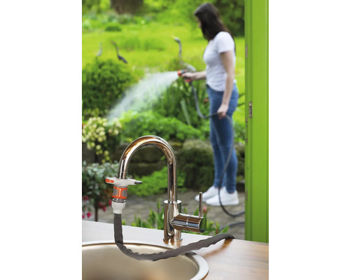 Trädgårdsscen med kran och ansluten trädgårdsslang, i bakgrunden en kvinna som vattnar växter