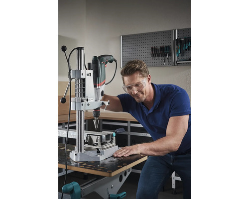 En man med skyddsglasögon använder en bordsborrmaskin i en verkstad.