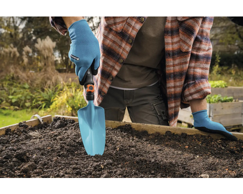 Person med trädgårdshandskar använder en trädgårdsspade i en upphöjd rabatt.