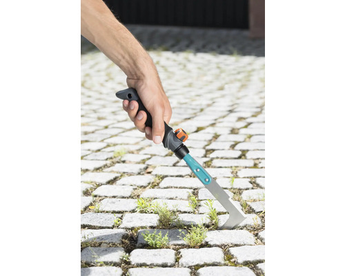 Hand håller en fogskrapa för att ta bort ogräs mellan gatstenar.