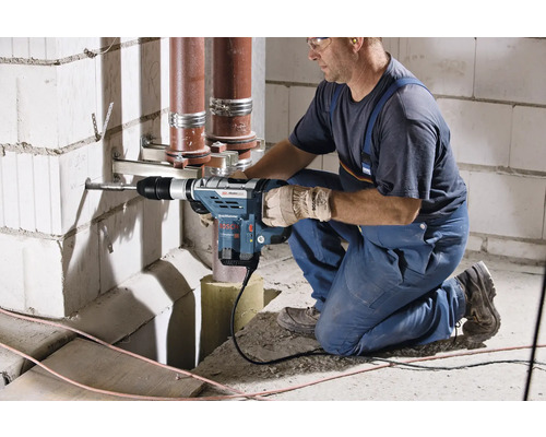 En hantverkare borrar ett hål i en vägg med en borrhammare, rör löper i bakgrunden.