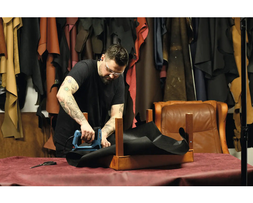 En hantverkare fäster med en elektrisk häftpistol en svart tygbit på en träram i en verkstad.