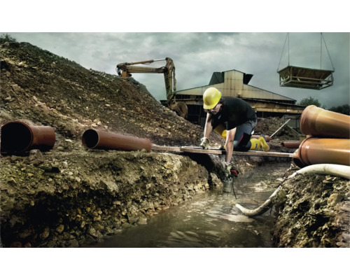 Byggnadsarbetare som arbetar i ett dike med rör, grävmaskin och kran i bakgrunden.