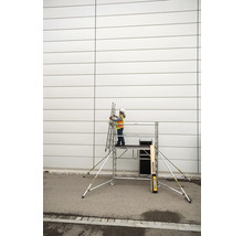 En person med skyddshjälm och säkerhetsväst står på en rullställning och håller en stege mot en vägg.