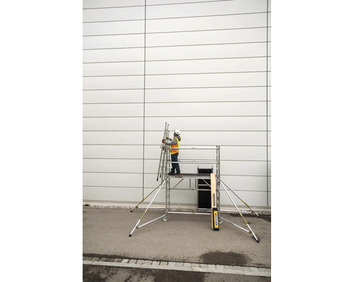 En person med skyddshjälm och säkerhetsväst står på en rullställning och håller en stege mot en vägg.