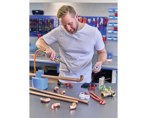 En man löder ett kopparrör med en brännare medan han håller i lödtenn. Diverse verktyg och rördelar av koppar ligger på bordet.