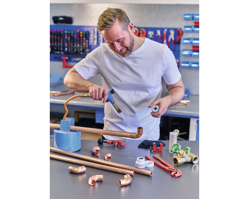 En man löder kopparrör med en lödkolv i en verkstad. Olika verktyg och rör ligger på bordet.