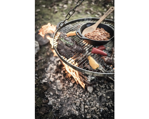Hängande grill över öppen eld med biff, grönsaker och bönor i en panna.