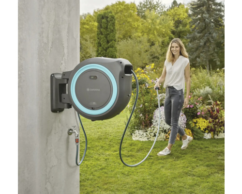 Trädgårdsscen med slangbox för trädgårdsbevattning, monterad på en vägg. En kvinna håller änden av slangen.