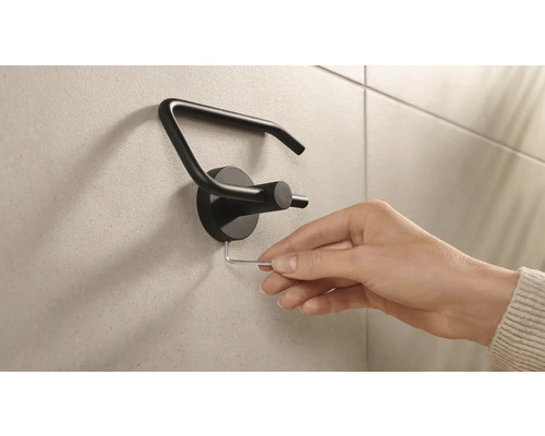 En hand monterar en svart toalettpappershållare på en vägg med en insexnyckel.